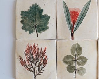 May include: Four square ceramic tiles with an off-white base, each featuring a distinct botanical print. The designs include a dark green maple-like leaf, a red flower with green leaves, a reddish-brown coniferous sprig, and a green rose-like leaf.