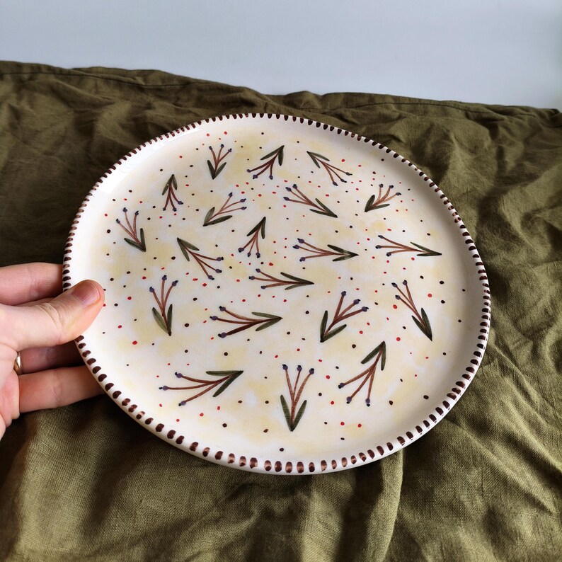 May include: A white ceramic plate with a hand-painted design of small green and brown sprigs with purple flowers and red dots on a yellow background. The plate has a brown rim.
