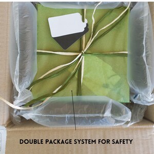 May include: A brown cardboard box with a green and white gift wrapped inside. The gift is wrapped in tissue paper and tied with a ribbon. The box is filled with air-filled packaging to protect the gift during shipping. The text "DOUBLE PACKAGE SYSTEM FOR SAFETY" is printed on the box.