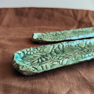 May include: Two handmade, rectangular ceramic trays with a botanical leaf and berry design. The trays are a mottled teal green color and sit on a brown surface. The trays are ideal for holding jewelry or small items.