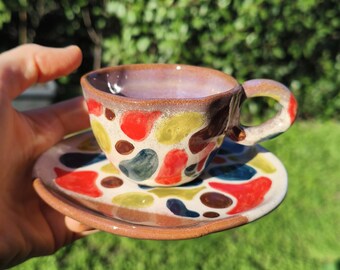 May include: A handmade ceramic teacup and saucer set. The cup and saucer are decorated with colorful, abstract shapes in red, yellow, blue, and brown against a cream background. The cup has a brown handle and rim.