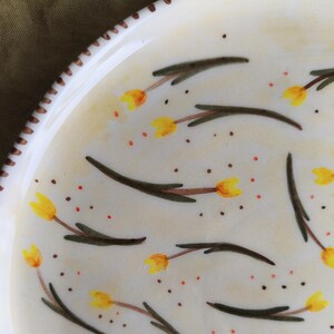 May include: A white ceramic plate with a brown rim features a hand-painted design of yellow flowers with green stems and brown dots on a cream background.