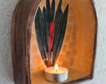 Può includere: Un portacandele da parete in ceramica con cornice ad arco, esterno marrone scuro e interno giallo. Il pannello posteriore presenta un disegno di foglie verdi dipinte con un accento rosso centrale e contiene una piccola candela bianca.
