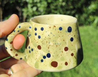 May include: A small, handmade ceramic mug with a light yellow-green base color. The mug is decorated with blue and brown polka dots. The handle is a matching light yellow-green color. The mug is held in a person's hand.