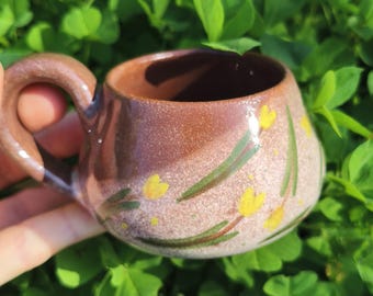 Op de afbeelding: Een kleine, handgemaakte keramische mok met een bruine buitenkant en een donkerbruine binnenkant. De mok heeft een gebogen handvat en is versierd met gele bloemen en groene bladeren. De mok heeft een diameter van ongeveer 7,6 cm.