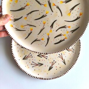 May include: Two ceramic plates with a hand-painted design of yellow flowers and green stems on a cream background. The smaller plate is stacked on top of the larger plate.