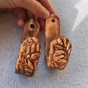 Op de afbeelding: Vier bruine keramische wandhaken met een bladeren design. De haken zijn rechthoekig met een gat aan de bovenkant om op te hangen. De onderkant van de haken heeft een gedetailleerd reliëf van bladeren en andere botanische elementen.