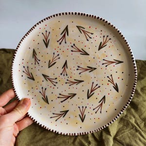 May include: A white ceramic plate with a hand-painted design of green and brown sprigs with purple and red dots. The plate has a brown rim with a decorative pattern.