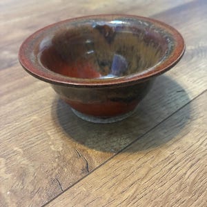 Könnte beinhalten: Eine handgefertigte Keramikschale mit glänzendem Finish. Die Schale hat einen rotbraunen Rand, der in eine Mischung aus braunen und orangenen Farbtönen übergeht. Der Innenraum hat ein dunkles, glänzendes Aussehen. Die Schale steht auf einem hellen Sockel.