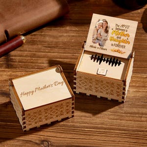 May include: Two light-coloured wooden music boxes with laser-cut patterns. One box is closed with the text Happy Mother's Day. The other is open, displaying a photo of two people and the text The LOVE between a Mother and Daughter is FOREVER.