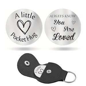 May include: Two silver pocket hug tokens with black text and heart designs. One says "A little Pocket Hug" and the other says "Always Know You Are Loved." A black leatherette pouch holds one token, with a keyring attached.