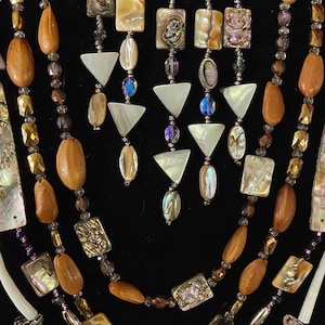Puede incluir: Collar multicadena con cuentas de concha de abulón iridiscente, madera y cristal. El collar tiene un diseño en capas y en cascada con una variedad de formas y tamaños de cuentas.