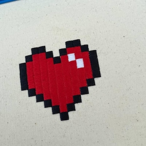 以下が含まれることがあります： 白い布地に赤いピクセルで刺繍されたハート。ハートは黒い輪郭で囲まれています。