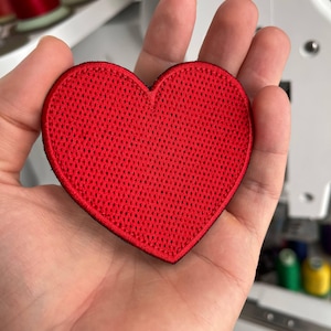 Pode incluir: Um patch em forma de coração vermelho brilhante com uma superfície texturizada semelhante a uma malha. O patch tem uma borda preta e é segurado em uma mão. O fundo mostra bobinas de linha e uma máquina de costura.