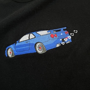 Peut inclure: Un t-shirt noir avec une voiture brodée en bleu. La voiture a un aileron et le texte "GT-R" est brodé à l'arrière.