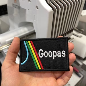 Puede incluir: Parche rectangular negro con la palabra "Goopas" en blanco y un diseño colorido de líneas azules, amarillas, verdes y rojas. El parche se sostiene en una mano, con una máquina de bordar en el fondo.