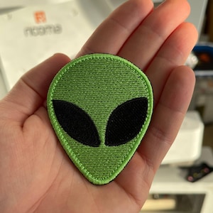 Könnte beinhalten: Ein grüner, gestickter Aufnäher in Form eines Alien-Kopfes mit großen, schwarzen, ovalen Augen. Der Aufnäher wird in einer Hand gehalten. Ein lustiges Accessoire.