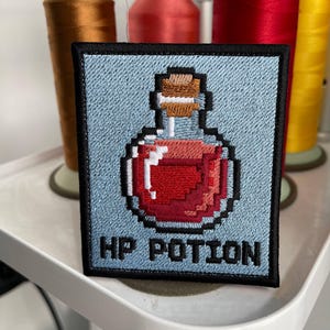 Puede incluir: Parche bordado cuadrado con una botella de poción roja pixelada con tapón de corcho sobre fondo azul claro. El texto "HP POTION" está debajo. El parche tiene un borde negro. Para los aficionados a los videojuegos.