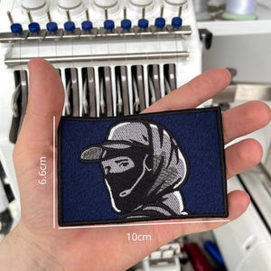 Può includere: Toppa ricamata blu navy con un disegno di un volto stilizzato con berretto e copertura per il viso. La toppa misura 10 cm per 6,6 cm. Il design è in tonalità di grigio e nero, con un bordo nero.