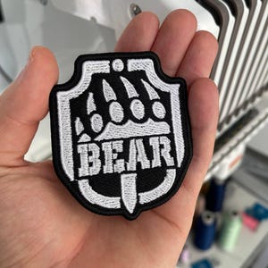 May include: A black and white embroidered patch in a shield shape. The patch features a bear claw design above the word "BEAR" in white block letters. The patch is held in a hand.