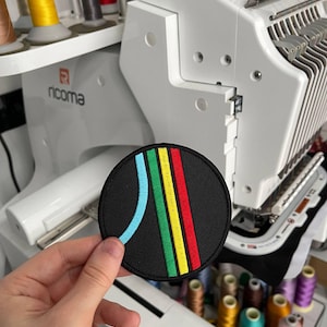 May include: A black, circular embroidered patch with a retro design. It features a blue arc and three vertical stripes in green, yellow, and red. The patch is held in front of an embroidery machine.