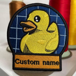Puede incluir: Parche bordado con un diseño de pato amarillo dentro de un círculo de cuadrícula azul. El parche tiene un borde negro y una sección rectangular en la parte inferior con el texto "Custom name" en marrón. El fondo incluye carretes de hilo.