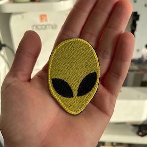 Op de afbeelding: Geel geborduurde alien patch met zwarte ogen en een zwarte rand. De patch wordt in een hand gehouden. De achtergrond omvat spoelen garen en een naaimachine.