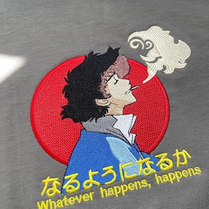 Puede incluir: Camiseta gris con un bordado de un personaje de anime. El personaje tiene pelo negro, una chaqueta azul y un fondo de círculo rojo. El texto "Whatever happens, happens" está bordado en amarillo.