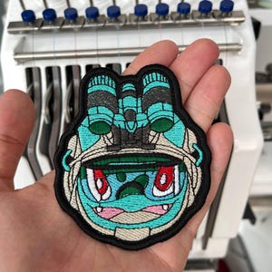 Puede incluir: Parche bordado con un personaje de dibujos animados con cara turquesa, ojos rojos y contorno negro. El parche tiene un borde negro y es para ropa o accesorios.