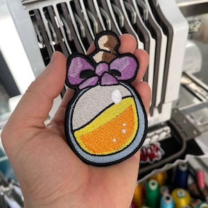 Könnte beinhalten: Gestickter Aufnäher mit einem Fläschchen-Design. Der Aufnäher hat eine lila Schleife, einen braunen Verschluss und eine Flasche, die mit gelber und orangefarbener Flüssigkeit gefüllt ist. Der Aufnäher ist schwarz und blau umrandet.