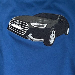 以下が含まれることがあります： 青いスウェットシャツ。黒い車の刺繍デザインが施されています。車は詳細なフロントグリル、ヘッドライト、ホイールを備えています。アウディのロゴが見えます。スウェットシャツは柔らかく快適な素材で作られています。車のデザインは約30cmです。