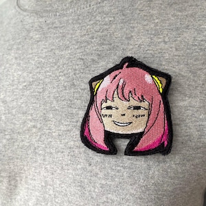 May include: Embroidered patch featuring a smiling anime character with pink hair and yellow accents. The patch is outlined in black and is attached to a gray t-shirt. The character's expression is cheerful.