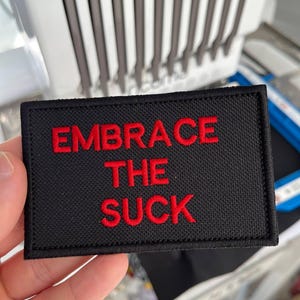 May include: Black rectangular patch with the words "EMBRACE THE SUCK" embroidered in red. The patch is held in a hand, with a blurred background of a machine.