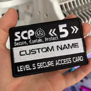 Op de afbeelding: Zwart en wit geborduurde patch met de tekst "SCP Secure, Contain, Protect" en "CUSTOM NAME". De patch heeft ook het nummer "5" en de woorden "LEVEL 5 SECURE ACCESS CARD".