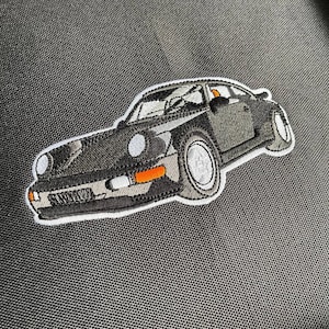 Puede incluir: Un parche bordado que representa un coche deportivo clásico de color negro con detalles blancos y acentos naranjas.