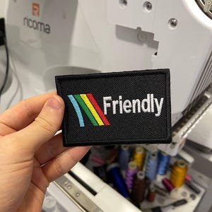 Könnte beinhalten: Ein schwarzer rechteckiger Aufnäher mit dem Wort "Friendly" in Weiß und einem farbenfrohen Streifendesign. Der Aufnäher wird vor einer Stickmaschine gehalten. Das Design umfasst blaue, grüne, gelbe und rote Streifen.