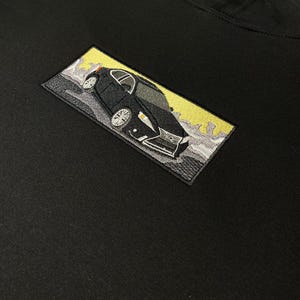 May include: Black hoodie with an embroidered image of a black car on a yellow background.