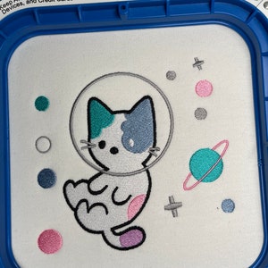 Puede incluir: Diseño bordado de un gato blanco con un casco espacial, rodeado de planetas y estrellas de colores. El diseño está en una tela blanca en un aro de bordado.