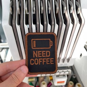 May include: A black rectangular patch with the words "NEED COFFEE" embroidered in brown thread. The patch has a brown border and a battery icon. The background shows an embroidery machine and spools of thread.