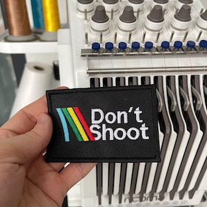 Könnte beinhalten: Schwarzer rechteckiger Aufnäher mit dem weißen Text "Don't Shoot". Der Aufnäher hat ein farbenfrohes Design aus blauen, grünen, gelben und roten Streifen. Der Aufnäher wird vor einer Stickmaschine gehalten.
