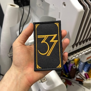 Può includere: Una toppa rettangolare nera con bordo dorato e il numero "33" in oro. La toppa è tenuta in mano, con una macchina da ricamo sullo sfondo. Il numero "33" è stilizzato con un carattere in grassetto.
