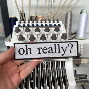 Puede incluir: Parche bordado con la frase "oh really?" en negro sobre fondo blanco, que mide 15 cm de largo y 3,8 cm de ancho. El parche se sostiene frente a una máquina de bordar.