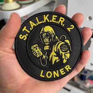 Op de afbeelding: Een ronde, zwarte geborduurde patch met gele tekst en een gele afbeelding van een persoon met een gasmasker. De tekst luidt "S.T.A.L.K.E.R. 2 LONER". De patch is bedoeld voor jassen of tassen.