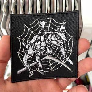 Op de afbeelding: Vierkante zwarte patch met een wit geborduurd ontwerp van een figuur gevangen in een spinnenweb. De patch heeft een zwarte rand en wordt in een hand gehouden.