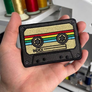 May include: Embroidered patch in the shape of a retro cassette tape. The patch features a tan background with colorful stripes of red, yellow, green, and blue. The cassette tape is outlined in black, with the word "INDEX" visible.