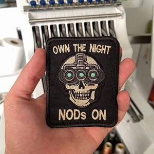 Può includere: Toppa ricamata nera con la scritta "OWN THE NIGHT" e "NODS ON". La toppa presenta un teschio con occhiali per la visione notturna con lenti verdi. La toppa è quadrata e ha un bordo nero.