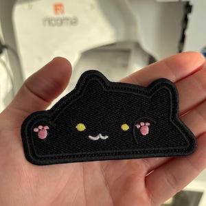 May include: Black embroidered cat patch with yellow eyes, pink paw pads, and a white mouth. The patch is held in a hand, with a blurred background of a sewing machine.