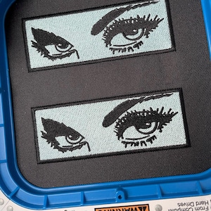 May include: Two embroidered patches featuring stylized black eyes with long lashes on a light blue background. The patches are rectangular and set against a black fabric, held within a blue embroidery hoop.