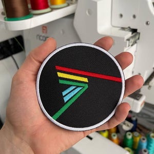 Könnte beinhalten: Ein runder, schwarzer, gestickter Aufnäher mit weißem Rand. Der Aufnäher zeigt ein farbenfrohes geometrisches Design mit roten, gelben, grünen und blauen Linien. Der Aufnäher wird in einer Hand gehalten, mit Garnrollen und einer Stickmaschine im Hintergrund.