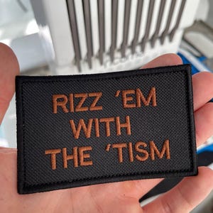 Könnte beinhalten: Schwarzer rechteckiger Aufnäher mit braun gesticktem Text "RIZZ 'EM WITH THE 'TISM". Der Aufnäher hat eine strukturierte Oberfläche.
